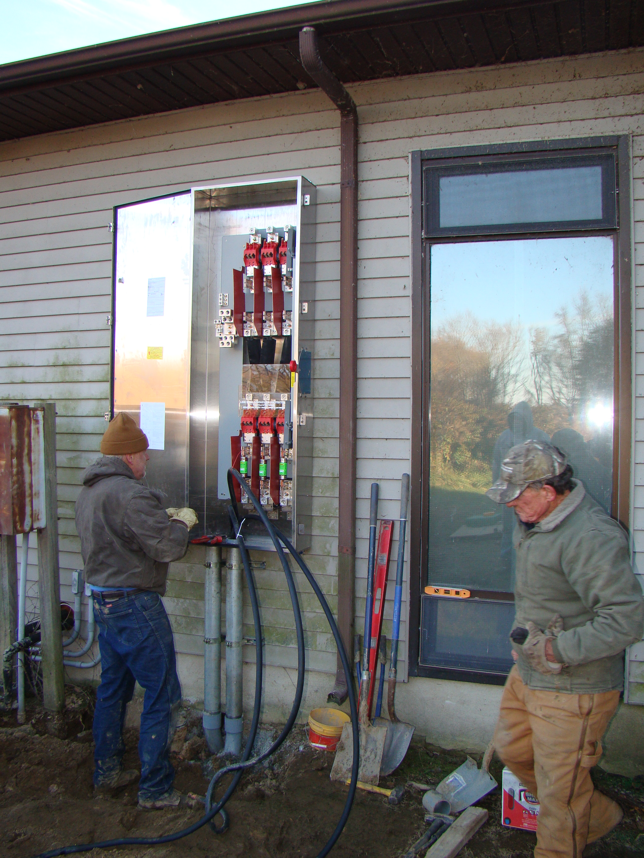 Two workers wiring a large electrical panel on the side of a house during installation work